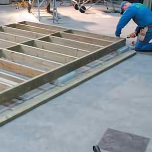 May include: A partially constructed wooden frame, likely for a deck or platform, with a grid-like structure. The wood is unfinished and light brown. A person in blue work clothes is working on the frame. The setting appears to be a construction site.