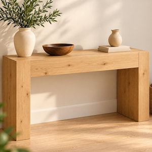 Puede incluir: Una mesa consola rectangular de roble claro con un patrón de veta de madera visible. Encima hay un jarrón de cerámica beige grande con follaje verde, un cuenco de madera marrón oscuro y un jarrón de cerámica beige pequeño sobre dos libros.