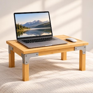 Puede incluir: Una mesa rectangular para portátil hecha de bambú con cuatro patas cortas y soportes de esquina de metal plateado. Un portátil plateado está abierto sobre la superficie mostrando una imagen de montañas y un lago, con un ratón blanco a la derecha.