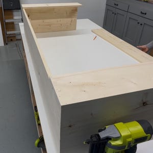 May include: A wooden frame is being assembled with a nail gun. The frame is made of unfinished pine wood and is being built on a white surface.