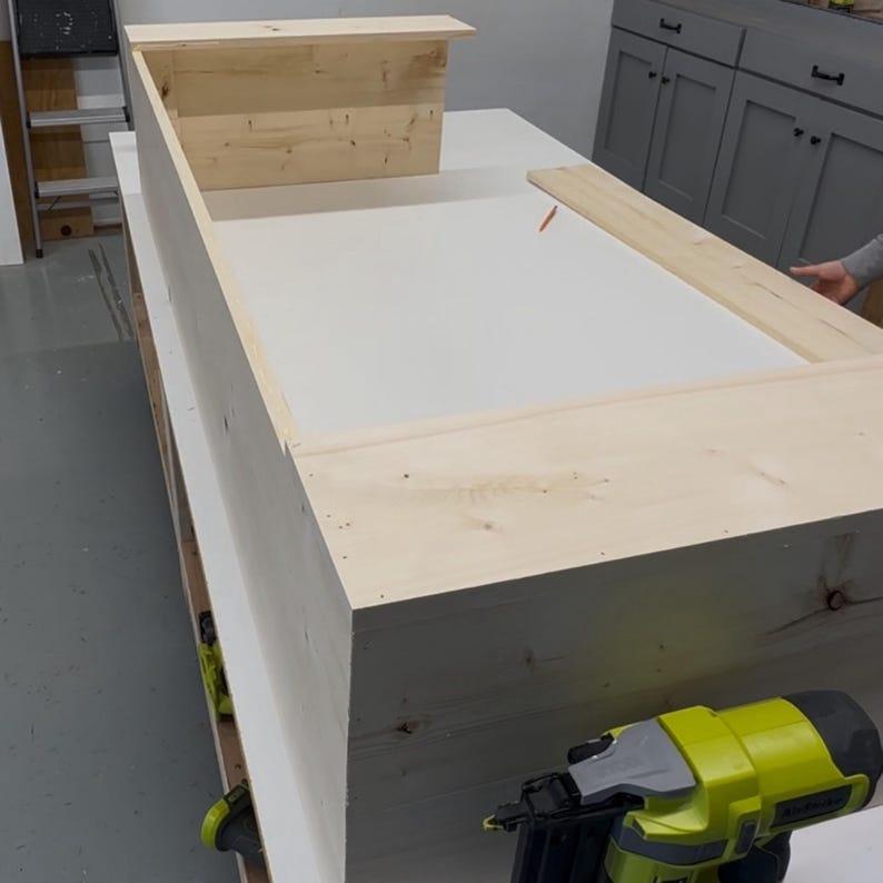 May include: A wooden frame is being assembled with a nail gun. The frame is made of unfinished pine wood and is being built on a white surface.