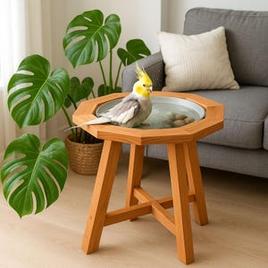 May include: A yellow and grey cockatiel bird is taking a bath in a round wooden bird bath on a wooden table. The bird bath is filled with water and has a few small stones in it. The bird is standing in the water and is looking at the camera.
