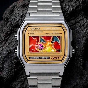 Puede incluir: Un reloj digital plateado con una esfera dorada que presenta un gráfico de un dragón naranja lanzando fuego. El marco de la pantalla incluye los textos CASIO, ALARM CHRONO, WATER RESIST y WR. El brazalete es de eslabones metálicos.