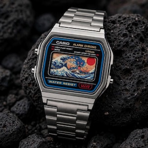 Könnte beinhalten: Eine silberfarbene Digitalarmbanduhr mit Metallgliederarmband. Das Zifferblatt zeigt den Text CASIO, ALARM CHRONO und WATER RESIST sowie eine grafische Illustration eines blauen Seedrachens in Wellen mit einer roten Sonne auf dem Display.