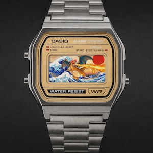 Könnte beinhalten: Eine silberfarbene Digitalarmbanduhr mit Metallgliederarmband und goldfarbenem Zifferblatt. Das Display zeigt eine Grafik eines drachenartigen Wesens, das Feuer über stilisierte blaue Wellen und eine rote Sonne speit, mit CASIO ALARM CHRONO.