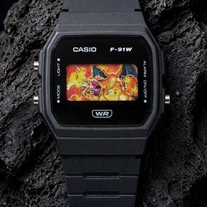 May include: A black digital wristwatch with a rectangular face featuring the text CASIO F-91W and WR. The display screen shows a colourful graphic of a fire-breathing dragon character. The watch has a black resin strap and side control buttons.