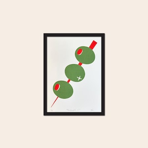 May include: A framed art print featuring a graphic illustration of three green olives on a red toothpick. The olives are arranged diagonally on a white background. The text "Stick with it" is printed at the bottom, along with a signature and edition number.