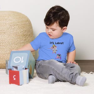 May include: A light blue toddler t-shirt featuring the text "It's Labrat!" above cartoon illustrations of a mouse and a purple creature. The child is wearing gray pants and white socks.