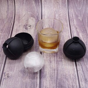 May include: Two black spherical silicone ice moulds, one open and one closed, alongside a clear glass tumbler containing a single large round ice sphere submerged in an amber liquid, with an additional loose ice sphere resting on the surface.