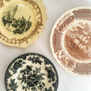 May include: Three ceramic plates with scalloped edges. One cream plate features a brown transferware print of Westminster Bridge and Big Ben. A second cream plate displays a brown ship scene labeled Canton Port 1830. A third plate has a black floral pattern.