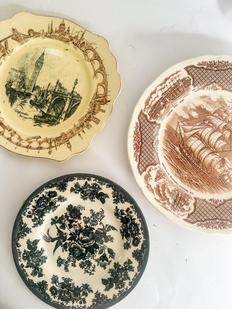 May include: Three ceramic plates with scalloped edges. One cream plate features a brown transferware print of Westminster Bridge and Big Ben. A second cream plate displays a brown ship scene labeled Canton Port 1830. A third plate has a black floral pattern.