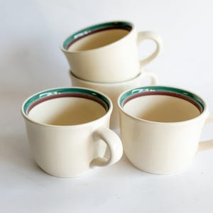 Può includere: Tazze in ceramica color crema con bordo verde scuro e marrone. Le tazze hanno una forma arrotondata con un manico curvo. Quattro tazze sono impilate, due in primo piano.