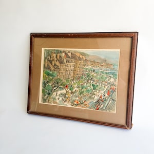 May include: Framed vintage print depicting a European cityscape with buildings, trees, and figures. The artwork features a brown wooden frame and a tan mat. The print showcases a colorful illustration of a town scene.
