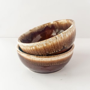 Peut inclure: Deux bols en céramique empilés, avec un glaçage marron et crème. Les bols ont une forme arrondie et une finition brillante. Le glaçage a un effet dégoulinant, créant un motif unique. Convient pour servir des aliments.
