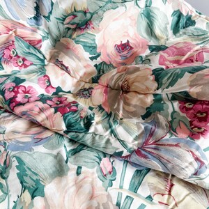 May include: A close-up of a floral patterned comforter. The comforter features large, soft pink, peach, and white flowers with green leaves against a white background. The fabric is slightly wrinkled, suggesting a soft, comfortable texture.