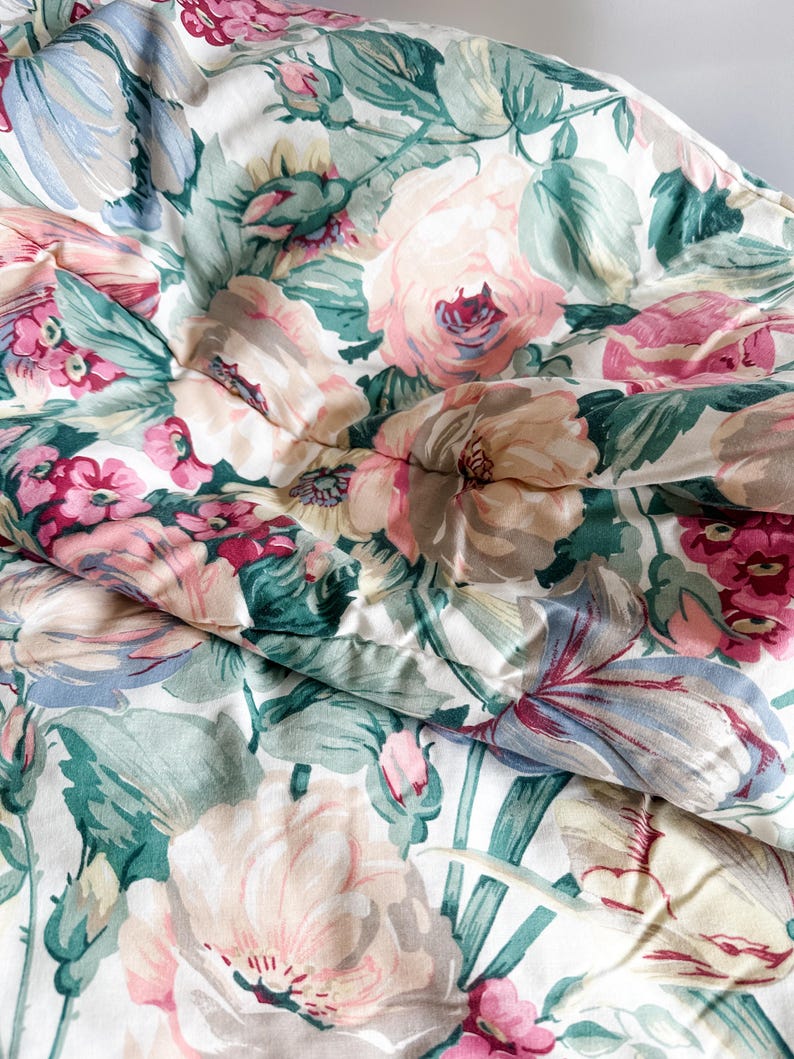 May include: A close-up of a floral patterned comforter. The comforter features large, soft pink, peach, and white flowers with green leaves against a white background. The fabric is slightly wrinkled, suggesting a soft, comfortable texture.