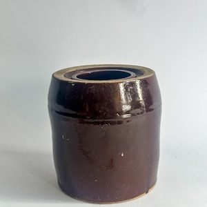 May include: A dark brown, cylindrical stoneware crock with a glossy finish. The antique container has a slightly flared rim and a recessed interior. The exterior shows subtle variations in color and texture, suggesting its age and use. A classic piece for kitchen or home decor.