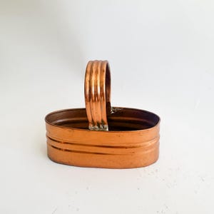 May include: A copper-colored, oval-shaped basket with a handle. The basket has a simple design with horizontal bands around the sides and a ribbed handle. The metal has a warm, reflective sheen.