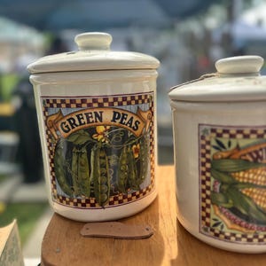 May include: Two cream-coloured ceramic canisters with lids. One is labelled "GREEN PEAS" and features a graphic of pea pods. The other has a corn graphic. They sit on a wooden surface, ideal for kitchen storage.