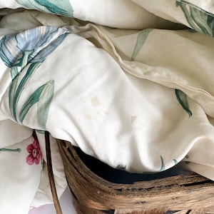 May include: A close-up of a white floral quilt with blue and green leaf and flower patterns. The quilt is partially folded, revealing a woven brown basket. The quilt has a soft, textured appearance.