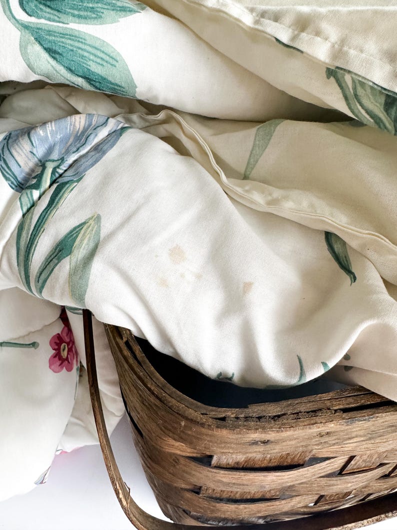 May include: A close-up of a white floral quilt with blue and green leaf and flower patterns. The quilt is partially folded, revealing a woven brown basket. The quilt has a soft, textured appearance.