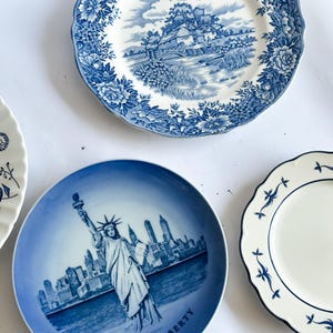 May include: Three ceramic plates with blue and white designs. One plate features a landscape with a bridge and cottage, another displays the Statue of Liberty and New York City skyline, and the third has a simple blue floral border pattern.