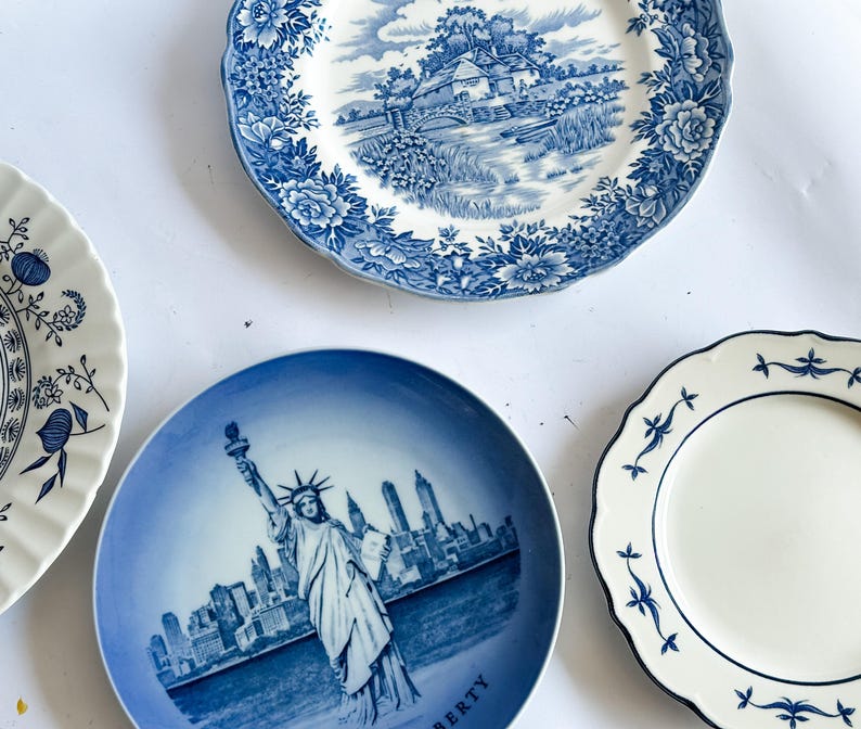 May include: Three ceramic plates with blue and white designs. One plate features a landscape with a bridge and cottage, another displays the Statue of Liberty and New York City skyline, and the third has a simple blue floral border pattern.