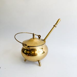 Könnte beinhalten: Ein kleiner, glänzender Messingkessel mit Deckel, Griff und drei kleinen Beinen. Der Kessel hat einen langen, abgewinkelten Ausguss mit einem Holzgriff. Der Deckel hat einen kleinen Griff und ist mit einem Scharnier befestigt. Der Kessel steht auf einer weißen Oberfläche.
