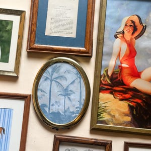 May include: A collection of framed art pieces, including a vintage print of a woman in a swimsuit, a poem, and a palm tree landscape. Frames vary in style, with wood and gold tones.