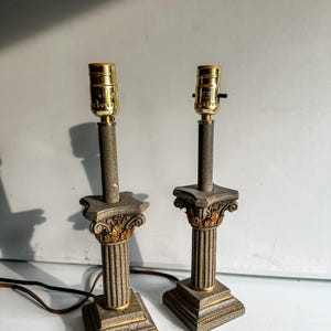 May include: Two decorative table lamps with a classical column design. The lamps have gold-coloured bulb sockets, grey columns with gold accents, and square bases. They are connected to a power source via a black cord.