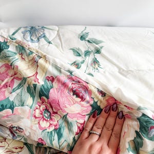 May include: A floral-patterned quilt featuring large pink roses, green leaves, and cream-colored accents. The quilt has a quilted design and a white background with a smaller floral pattern. The quilt is folded, revealing both sides of the fabric.