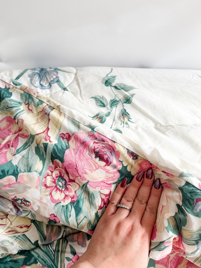 May include: A floral-patterned quilt featuring large pink roses, green leaves, and cream-colored accents. The quilt has a quilted design and a white background with a smaller floral pattern. The quilt is folded, revealing both sides of the fabric.