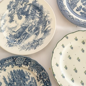 May include: Three ceramic plates are arranged together. One plate features a blue and white landscape scene with a bridge. Another plate displays a blue and white floral border with a rural scene. The third plate has a scalloped edge with green berries.