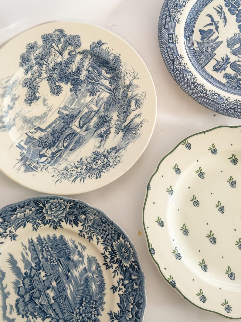 May include: Three ceramic plates are arranged together. One plate features a blue and white landscape scene with a bridge. Another plate displays a blue and white floral border with a rural scene. The third plate has a scalloped edge with green berries.