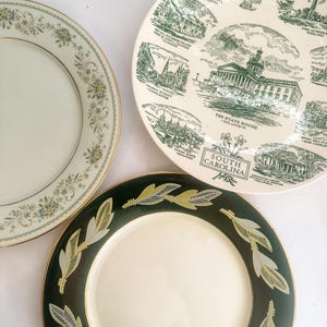 May include: Three ceramic plates: one with a floral border, one featuring green illustrations of South Carolina landmarks with text labels, and one with a dark green rim decorated with a repeating gold and white leaf pattern and gold trim.