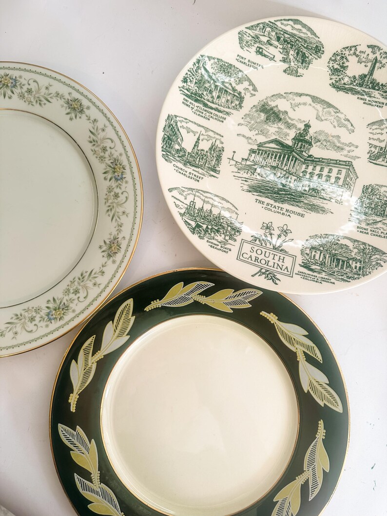 May include: Three ceramic plates: one with a floral border, one featuring green illustrations of South Carolina landmarks with text labels, and one with a dark green rim decorated with a repeating gold and white leaf pattern and gold trim.