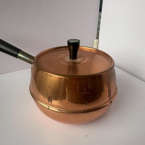 Könnte beinhalten: Ein Kupfertopf mit schwarzem Griff und Deckel. Der Topf hat eine abgerundete Form und eine glänzende Oberfläche.