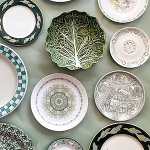 May include: A collection of ceramic tableware including a green cabbage-shaped bowl, a plate with a South Carolina state house print, a floral plate, a checkered plate, a Williamsburg Virginia plate, and several other plates with green and white patterns.