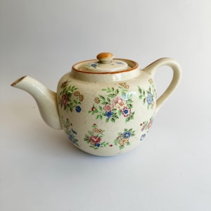 Puede incluir: Tetera de cerámica color crema con un estampado floral en rosa, azul y amarillo. La tetera tiene un pico curvo, un cuerpo redondeado y una tapa a juego con un pequeño pomo. El asa es curva y la tetera tiene una estética vintage.