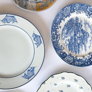 May include: A collection of four ceramic plates. One features a blue fish pattern on white, another displays a blue and white landscape scene with floral borders, a third shows a black and yellow landscape, and the fourth has a green-rimmed berry print.
