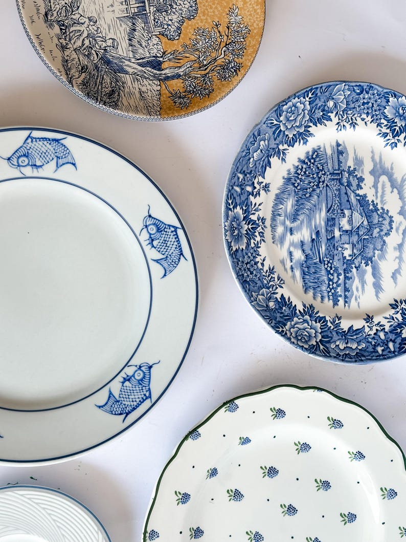 May include: A collection of four ceramic plates. One features a blue fish pattern on white, another displays a blue and white landscape scene with floral borders, a third shows a black and yellow landscape, and the fourth has a green-rimmed berry print.