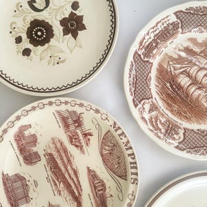 May include: Four ceramic plates with varied designs. One features brown floral patterns, another a brown ship illustration labeled Canton Port 1830, a third displays Memphis landmarks with text, and the fourth shows a partial orange floral motif.
