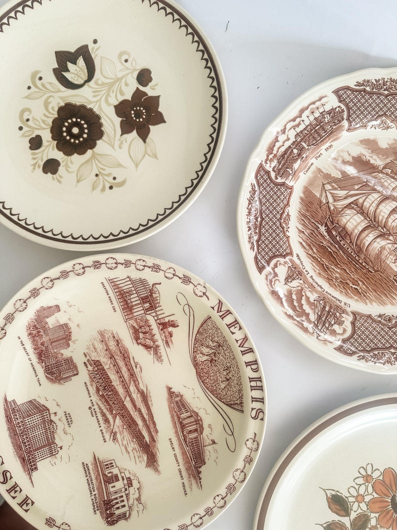 May include: Four ceramic plates with varied designs. One features brown floral patterns, another a brown ship illustration labeled Canton Port 1830, a third displays Memphis landmarks with text, and the fourth shows a partial orange floral motif.