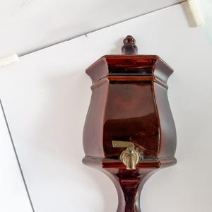 Puede incluir: Un dispensador de agua de cerámica marrón oscuro con un grifo de latón. El dispensador tiene una parte superior hexagonal y una base decorativa alargada. Diseñado para montaje en pared.