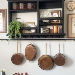 Puede incluir: Un estante de pared de madera negra con varios compartimentos que contienen tazas de vidrio rosa, una planta verde, una lata de cacao Ghirardelli y arte enmarcado. Debajo del estante, una barra negra sostiene cuatro sartenes de cobre.