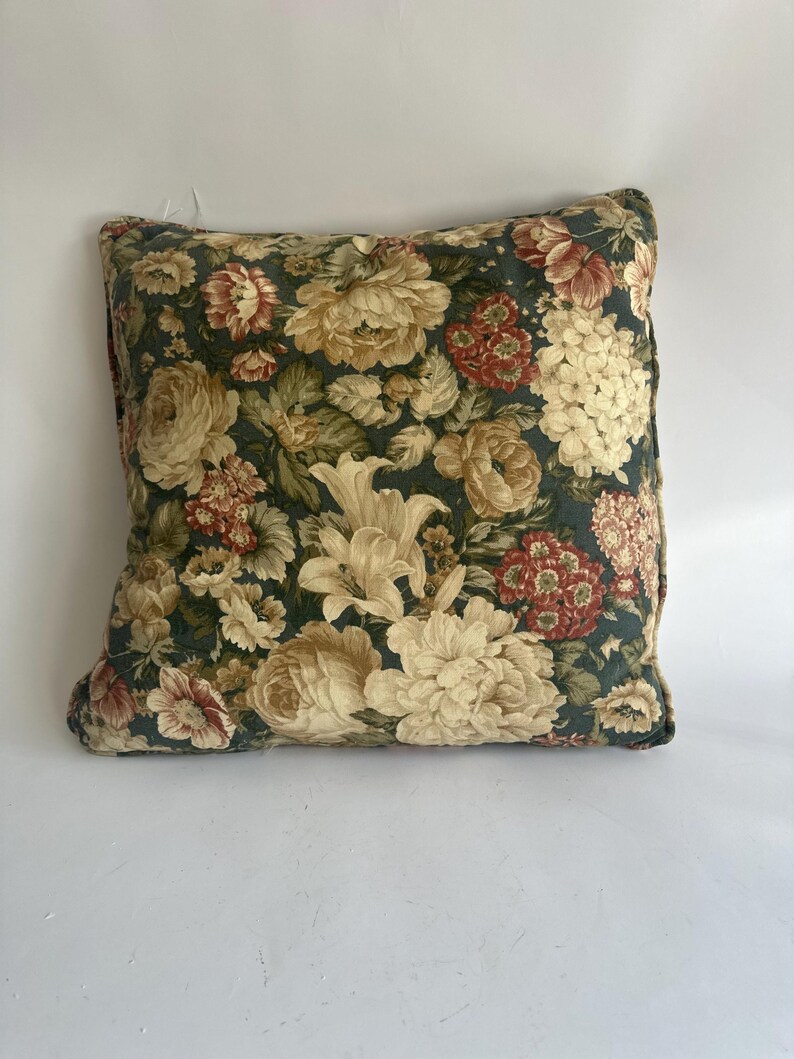 K&ouml;nnte beinhalten: Quadratisches Zierkissen mit dunkelgr&uuml;nem Hintergrund und gro&szlig;em Blumenmuster. Die Blumen sind in Creme-, Rot- und Gelbt&ouml;nen gehalten. Das Kissen hat ein weiches, strukturiertes Aussehen, was auf ein komfortables Design hindeutet.