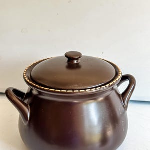 Peut inclure: Un pot en céramique brun foncé avec un couvercle assorti. Le pot a deux poignées latérales et une bande décorative autour du bord. Le couvercle a un petit bouton sur le dessus. Cet article de cuisine est adapté à la cuisson et au service.