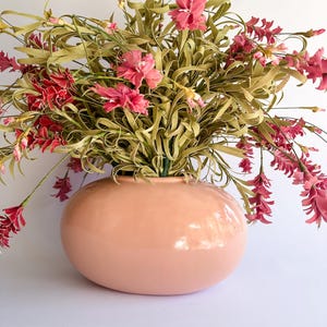 May include: A peach-colored, rounded ceramic vase filled with faux flowers. The arrangement features green, leafy stems and clusters of pink and red blossoms. The vase has a glossy finish and sits on a white surface.