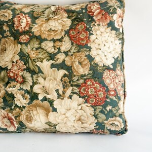 K&ouml;nnte beinhalten: Quadratisches Zierkissen mit dunkelgr&uuml;nem Hintergrund und floralem Muster. Das Muster umfasst gro&szlig;e cremefarbene, beige und rote Blumen mit gr&uuml;nen Bl&auml;ttern. Das Kissen hat einen dunkelgr&uuml;nen Rand.
