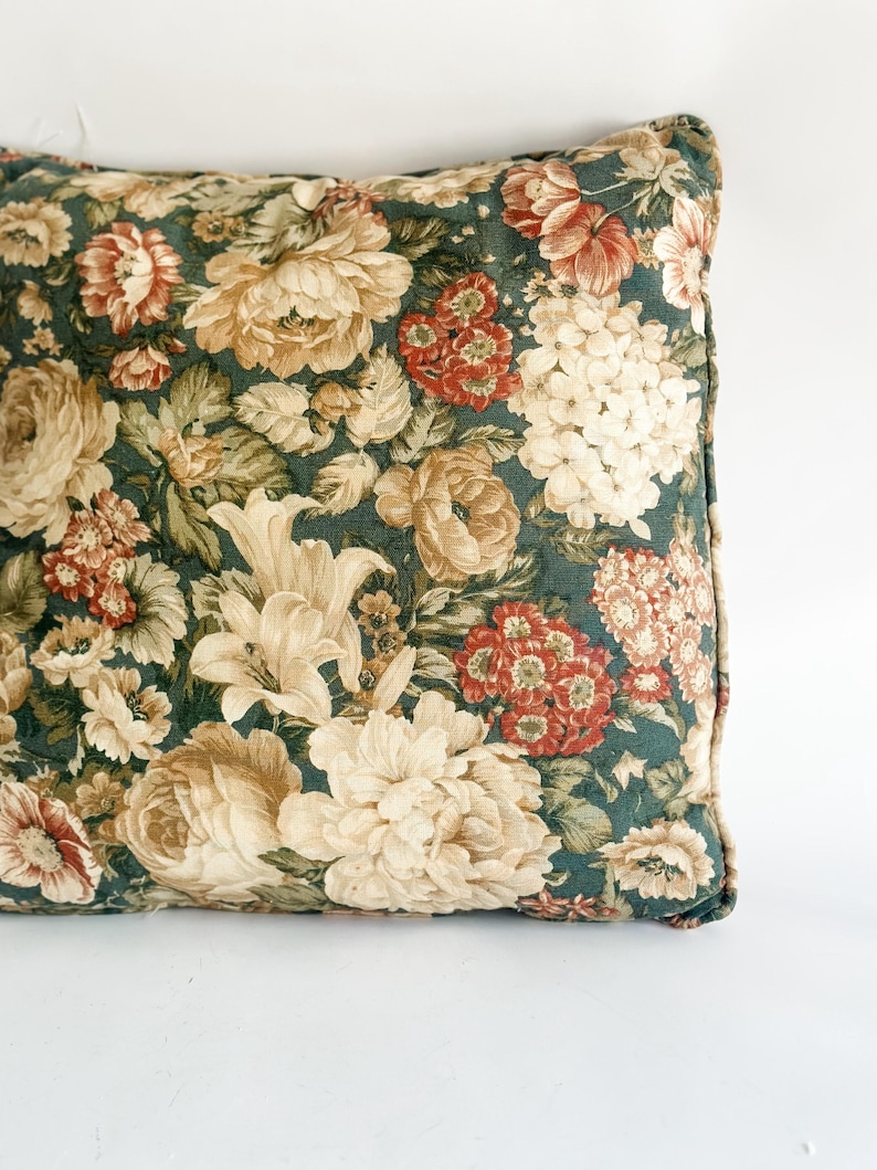 K&ouml;nnte beinhalten: Quadratisches Zierkissen mit dunkelgr&uuml;nem Hintergrund und floralem Muster. Das Muster umfasst gro&szlig;e cremefarbene, beige und rote Blumen mit gr&uuml;nen Bl&auml;ttern. Das Kissen hat einen dunkelgr&uuml;nen Rand.